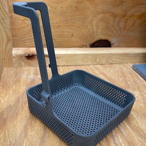 May include: A dark gray, rectangular basket with a handle. The basket has a perforated bottom and sides. The handle is attached to the side of the basket with a hinge. The basket is sitting on a wooden surface.