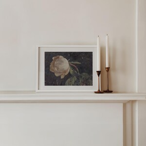 A moody floral painting of a white rose on a black background that goes with e a cottagecore or dark botanical and dark academia aethetic. The painting sits on a mantle.