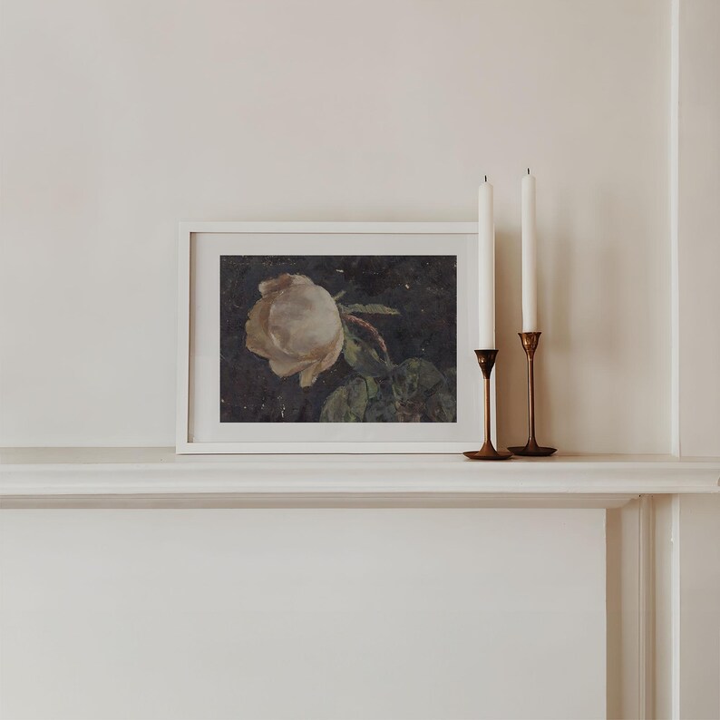 A moody floral painting of a white rose on a black background that goes with e a cottagecore or dark botanical and dark academia aethetic. The painting sits on a mantle.