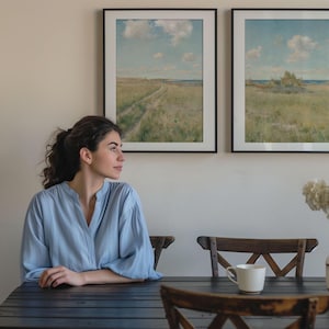 May include: A woman sits at a wooden table with two chairs. There are two framed landscape paintings on the wall behind her. A vase with white flowers sits on the table next to a white coffee mug.