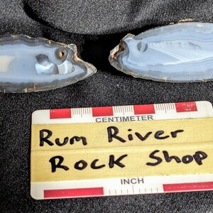 May include: Two agate slices with a white and gray banded pattern. The slices are on a black fabric background with a yellow tag that reads "Rum River Rock Shop".