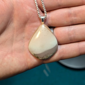 May include: A teardrop-shaped pendant made of white agate with brown and green inclusions. The pendant is attached to a silver chain.