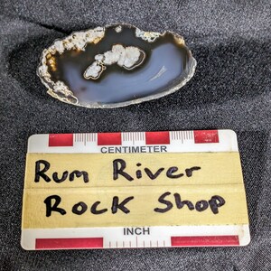 May include: A close-up of a gray and white agate slice with a brown border. The agate slice is about 2 inches in diameter. The slice is sitting on a gray fabric surface. A yellow label with black text reads "Rum River Rock Shop".