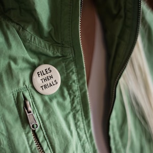 Könnte beinhalten: Hellgrüne Jacke mit silbernem Reißverschluss und einem weißen Knopf, der vorne angesteckt ist. Der Knopf trägt die Worte "FILES THEN TRIALS" in schwarzem Text.