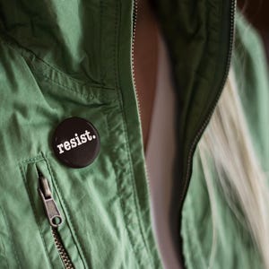 Puede incluir: Un botón marrón oscuro con la palabra "resist" en letras blancas está prendido a una chaqueta verde. La chaqueta tiene una cremallera y un bolsillo. El fondo está borroso, mostrando el pelo de una persona.