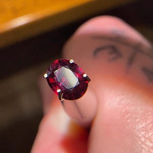 May include: A deep red oval gemstone set in a silver prong setting.
