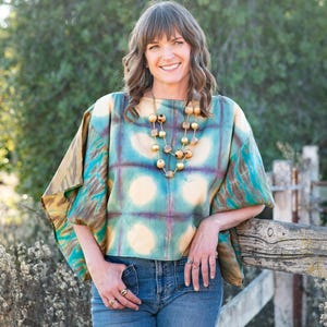 May include: A woman wearing a tie-dye top with a square pattern in shades of teal, blue, and yellow. She is also wearing blue jeans and a wooden bead necklace. The woman is smiling and leaning on a wooden fence.
