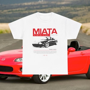May include: White t-shirt featuring a red "MIATA" graphic with Japanese text and a black car illustration. The shirt also includes the years "1989-1997" and additional Japanese text. A red convertible car is visible in the background.
