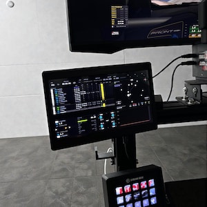 May include: A multi-screen racing simulator setup with a black frame. The screens display racing data and a race track view. A Stream Deck control panel is visible at the bottom, featuring multiple illuminated buttons.