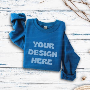 Puede incluir: Una sudadera de cuello redondo y manga larga en color azul real con el texto YOUR DESIGN HERE impreso en azul claro con una tipografía redondeada y negrita en el centro del pecho. La prenda cuenta con cuello, puños y dobladillo acanalados.