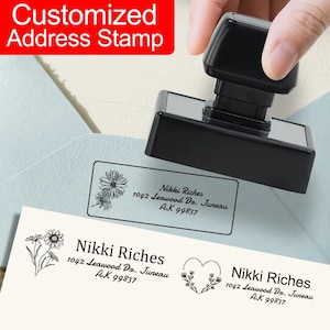 Puede incluir: Un sello de dirección negro personalizado con el texto "Customized Address Stamp" en un cuadro rojo. El sello se muestra en uso, imprimiendo una dirección con un diseño floral. La dirección dice "Nikki Riches, 1042 Leawood Dr. Juneau, AK 99837."