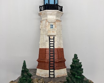 May include: A decorative lighthouse figurine with a black top and a blue light. The lighthouse has a white and brown body with a black ladder and a small window. Two green trees and a rocky base complete the scene.