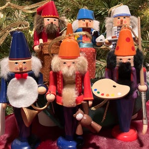 May include: A set of ten wooden nutcracker ornaments, each with a different profession and unique details. The ornaments are painted in various colors, including red, blue, green, and white. They are all wearing traditional nutcracker hats and have beards.