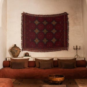 May include: A room featuring a long, low-profile rust-colored sofa with multiple brown throw pillows, a large dark red woven wall tapestry with a diamond pattern, a terracotta vase, a wooden bowl, and a floor cushion on a geometric rug.