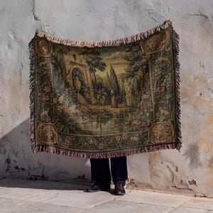 May include: A rectangular tapestry with a detailed landscape scene in shades of green, brown, and gold, fringed edges. The tapestry is held up against a weathered white wall, with a person's legs and feet visible below.