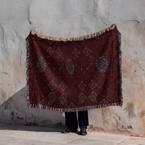May include: A rectangular woven throw blanket featuring a deep burgundy base color with intricate geometric patterns in shades of blue, cream, and black. The blanket has a decorative fringe along the top and bottom edges and a textured fabric finish.
