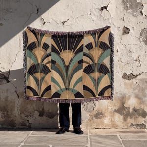 May include: A woven throw blanket featuring an Art Deco fan pattern in black, tan, and teal. The blanket has a fringed edge. The background is a weathered wall. The blanket is held up in front of a person.