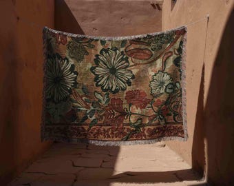May include: A rectangular woven throw blanket featuring a large-scale floral pattern in shades of deep red, olive green, and muted gold. The textile has a fringed edge along the bottom and sides, with a dense, intricate botanical design throughout.