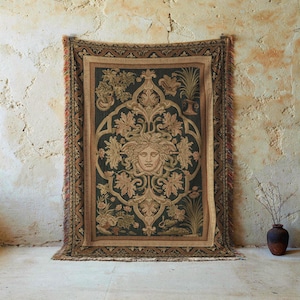 May include: A rectangular tapestry with a central Medusa face design, surrounded by floral and leaf motifs in shades of beige, brown, and green. The tapestry has a dark background and a fringed edge with red and orange accents. The tapestry is hanging on a textured wall.