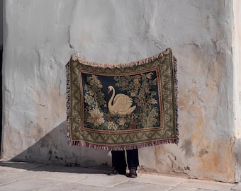 May include: A rectangular woven throw blanket featuring a central illustration of a white swan surrounded by detailed floral and botanical elements in shades of green, gold, and dark blue, finished with a decorative fringed border on all four sides.