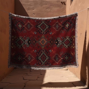 May include: A red patterned rug with a detailed geometric design hangs on a washing line. The rug has a fringe along the bottom edge. The background is a narrow alleyway with textured, light brown walls.