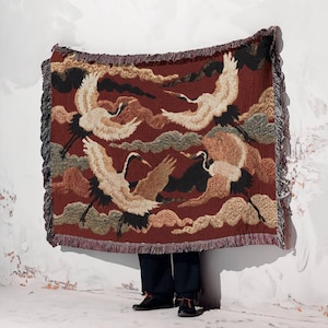 Puede incluir: Una manta rectangular con un fondo burdeos y un patrón de grullas y nubes volando. La manta tiene un borde con flecos en tonos marrones y grises. Las grullas están representadas en tonos blancos, marrones y negros.