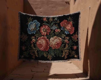 May include: A black tapestry with a floral design, featuring large pink, red, and blue flowers. The tapestry is fringed and hangs in an outdoor setting, with sunlight casting shadows on the walls.