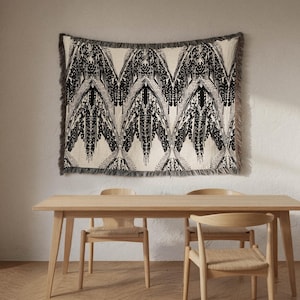 May include: A woven wall hanging with a black and white geometric pattern is displayed above a wooden dining table. The fringed edges of the wall hanging add texture. Two wooden chairs with woven seats are positioned on either side of the table.