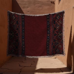 May include: A rectangular, fringed rug with a deep red central field and intricate patterned borders in navy, black, and touches of cream. The rug is suspended on a clothesline, showcasing its design and texture. The background features warm-toned walls.