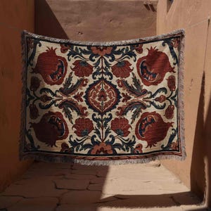 May include: A rectangular woven throw blanket with a detailed floral pattern. The design features large red pomegranate and floral motifs on a cream background, with accents of navy and olive green. The blanket has a fringed edge.