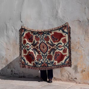 May include: A rectangular woven throw blanket featuring a symmetrical botanical pattern with large red pomegranate motifs, dark blue leafy vines, and cream-colored accents. The edges are finished with a decorative fringe on all four sides.
