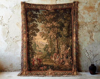 May include: A large tapestry depicting a pastoral scene with figures on horseback and in carriages, set within a forest. The tapestry features a detailed border and fringe, with a color palette of greens, browns, and reds. The tapestry is hanging on a textured, off-white wall.