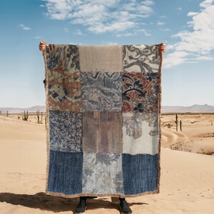 May include: A patchwork blanket in a variety of patterns and colours, including blue, brown, and beige. The rectangular blanket has a fringed edge. It is held up against a desert backdrop with a blue sky and some cacti.