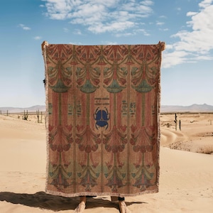 Puede incluir: Una manta rectangular con un diseño de inspiración egipcia. La manta presenta un patrón repetido de elementos florales estilizados en tonos de rojo, verde y azul sobre un fondo beige. Un gran escarabajo azul está centrado en la manta.