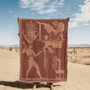 May include: A reddish-brown woven throw blanket with a classical Greek design. The design features figures, vases, and architectural elements in a lighter tan hue. The blanket has a fringed edge. The background is a desert landscape.