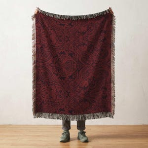 May include: A burgundy throw blanket with a dark paisley pattern and fringed edges. The blanket is held up, showcasing its intricate design and texture. The throw blanket is a home decor item.