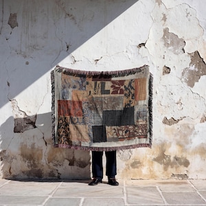 May include: A patchwork throw blanket with a fringed edge, featuring various patterned squares in shades of brown, black, and blue. The blanket is held up against a weathered white wall, with a person's legs and feet visible below.