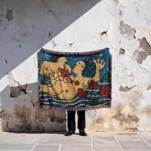 May include: A woven tapestry featuring a whimsical design with figures, flowers, and water elements in shades of blue, yellow, and red. The tapestry has a fringed edge and is held in front of a person against a textured white wall.
