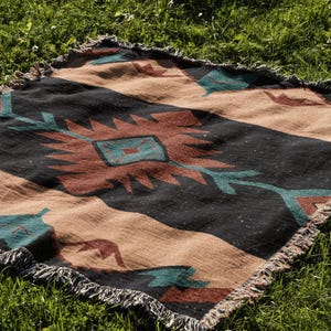 May include: A woven throw blanket with a southwestern pattern in shades of brown, black, and teal. The blanket has a fringed edge and is laid out on a bed of green grass.
