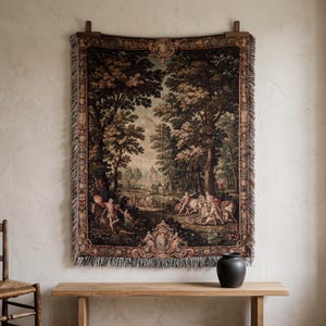 May include: A rectangular woven wall tapestry featuring a detailed landscape scene with tall trees, figures, and horses. The border is decorated with ornate scrollwork and floral motifs. The edges are finished with a fringed trim on all sides.