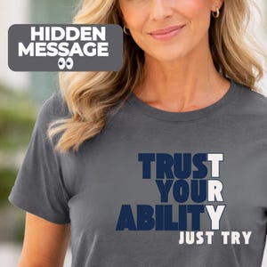 May include: Dark gray t-shirt with the words "TRUST YOUR ABILITY JUST TRY" in white and navy blue. The shirt also has a small graphic that says "HIDDEN MESSAGE". The shirt is made of a soft material.