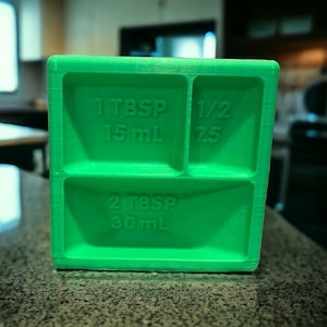 May include: Green plastic measuring tool with four compartments. The compartments are labeled with measurements in tablespoons and milliliters: 1 Tbsp 15 mL, 1/2 7.5, 2 Tbsp 30 mL.