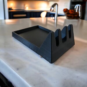 May include: Black utensil holder with a tray. The holder has three slots for utensils and the tray is made of a smooth, black material.