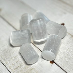 May include: A group of seven selenite crystal wands, each with a smooth, white, and translucent surface. The wands are arranged on a light wood surface.