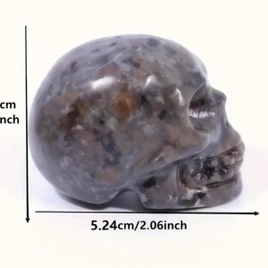May include: A dark gray and brown skull sculpture made of polished stone. The skull is approximately 1.51 inches tall and 2.06 inches wide.
