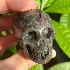 May include: A black and grey crystal skull with purple and white flecks. The skull is carved from a natural stone and has a detailed design.