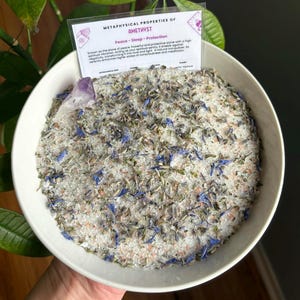 Könnte beinhalten: Eine weiße Schüssel gefüllt mit Badesalz, Lavendel und blauen Blütenblättern. Ein kleiner Amethystkristall liegt auf dem Salz. Eine Karte mit dem Text "METAPHYSISCHE EIGENSCHAFTEN VON AMETHYST Frieden - Schlaf - Schutz" ist im Hintergrund sichtbar.