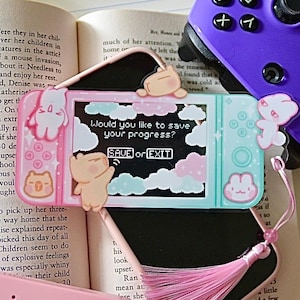 Può includere: Una Nintendo Switch Lite rosa pastello e blu con uno schermo che mostra un messaggio di salvataggio. La console è decorata con ciondoli a forma di animali e un nappa rosa. Un controller viola è visibile sullo sfondo.