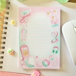 Puede incluir: Un bloc de notas rosa y verde con ilustraciones de tecnología retro, incluyendo una cinta de cassette, una cámara y un teléfono plegable. El bloc de notas tiene el texto "SnugglyAsh" en la parte inferior.