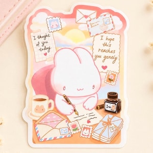 May include: A die-cut sticker featuring a white rabbit writing a letter with a fountain pen. The scene includes an ink bottle, a coffee mug, envelopes, and stamps. Text reads: I thought of you today. I hope this reaches you gently.